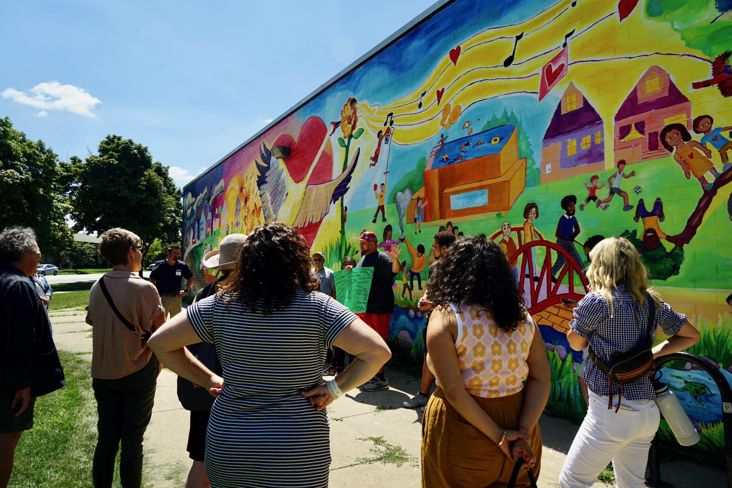 people facing away from the camera looking at a bright, colorful outdoor mural depicting community